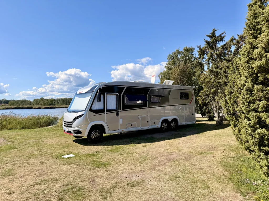 Der Dethleffs Globetrotter XLi steht auf der Wiese von Stellplatz 199, links das Wasser mit Schilfgürtel, rechts hohe Wacholderbäume.