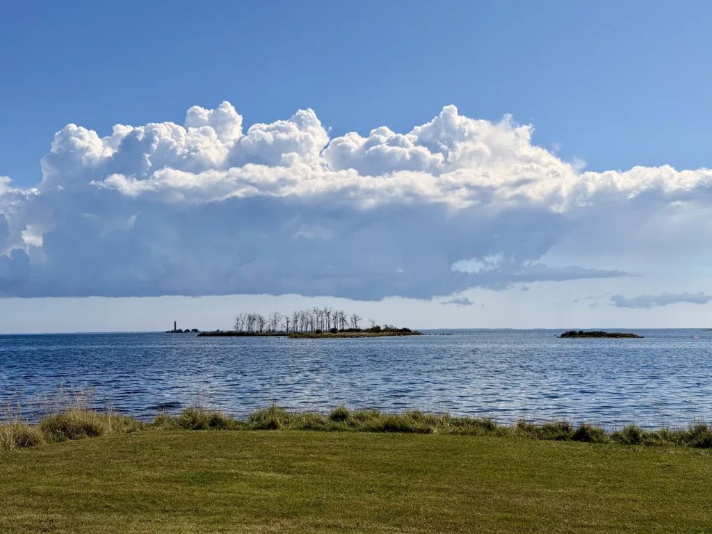 Blick von der Uferwiese über die Ostsee auf eine kleine bewaldete Schäreninsel, dahinter ein Leuchtturm und eine lange Wolkenbank.