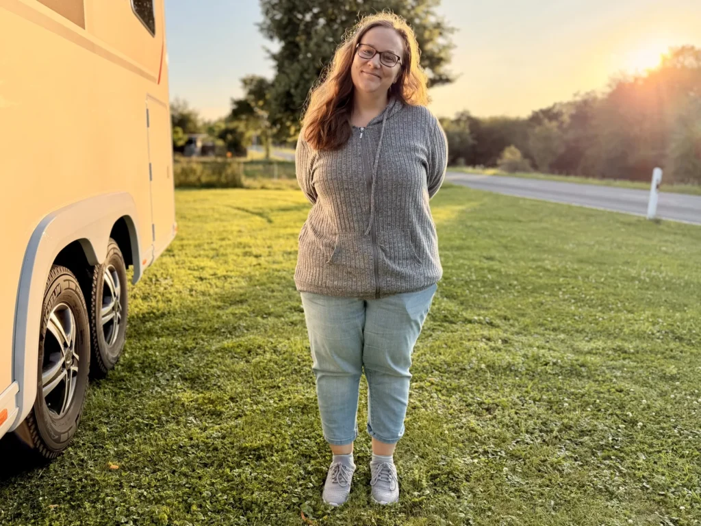 Eine Person steht neben dem Wohnmobil auf der Wiese, dahinter geht die Sonne über der Straße unter.