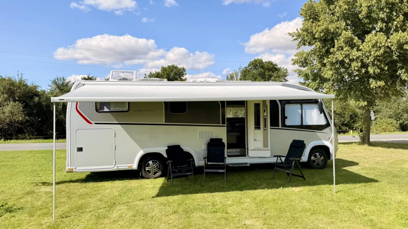 Unser Wohnmobil steht mit ausgefahrener Markise auf der Wiese, davor drei Campingstühle, dahinter eine große Eiche.
