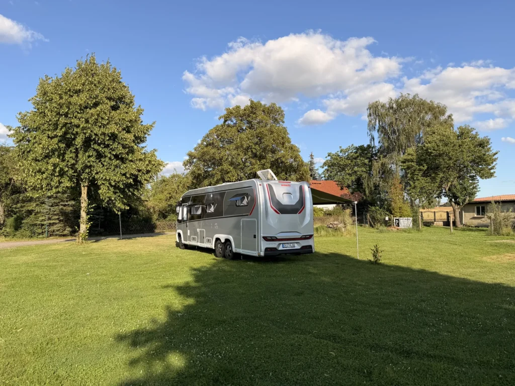 Unser Wohnmobil von schräg hinten auf der gemähten Wiese, im Hintergrund Bäume und die Dächer des Ortsrands.