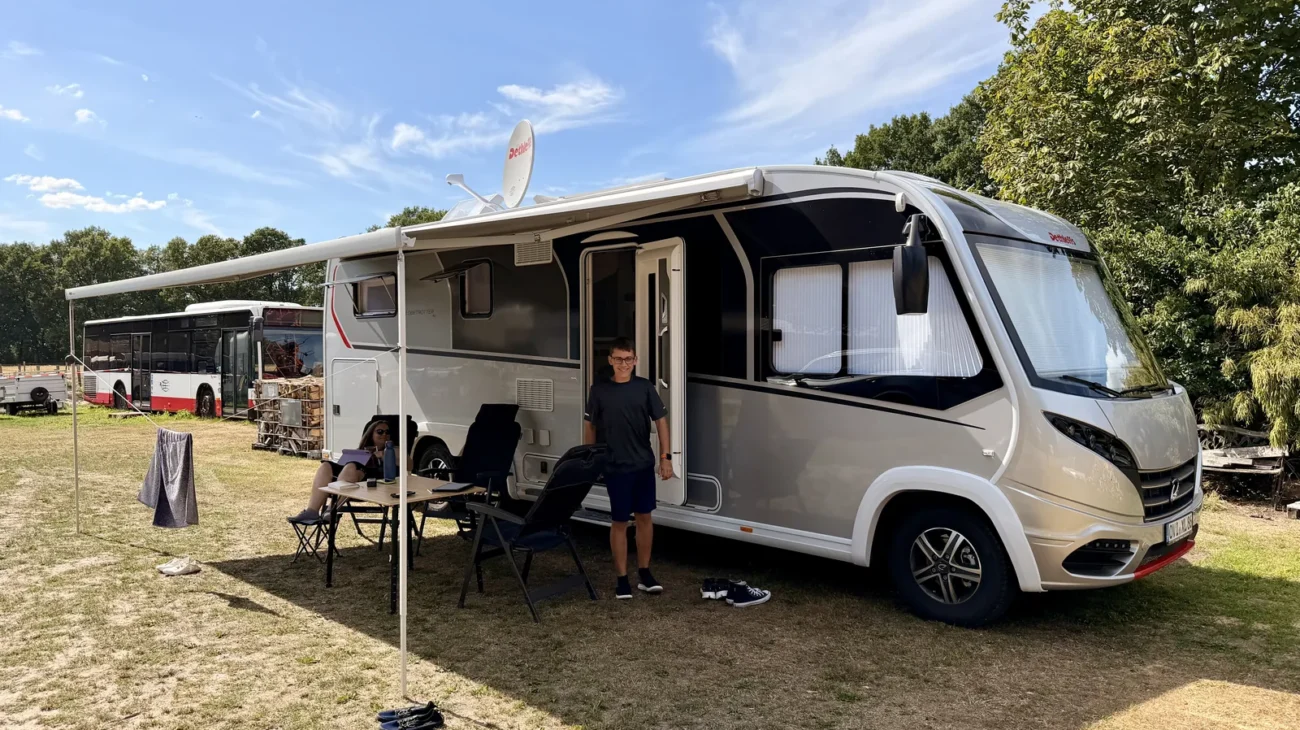 Unser Wohnmobil steht mit ausgefahrener Markise auf der Wiese, davor Campingtisch und Stühle, im Hintergrund ein alter Bus und Bäume.