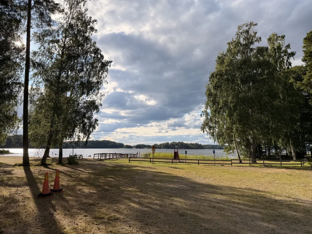 Der Badeplatz am See mit Holzsteg und Sprungturm, gesäumt von Birken, davor die Liegewiese.