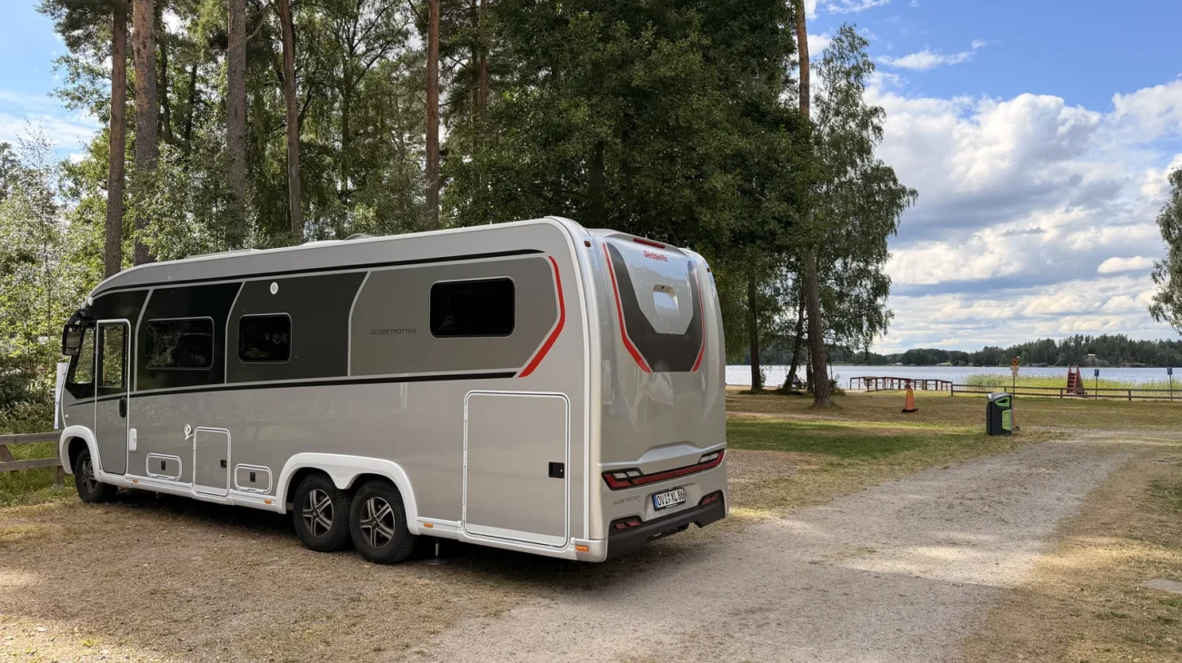 Unser Wohnmobil Luna steht auf dem Kiesplatz unter hohen Kiefern, im Hintergrund der See mit Badesteg.