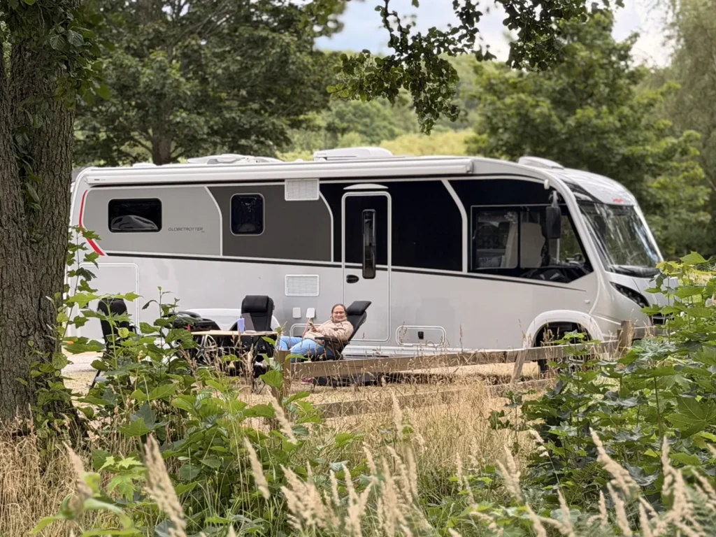 Blick durch hohes Gras auf unser Wohnmobil, davor sitzt eine Person am Campingtisch.