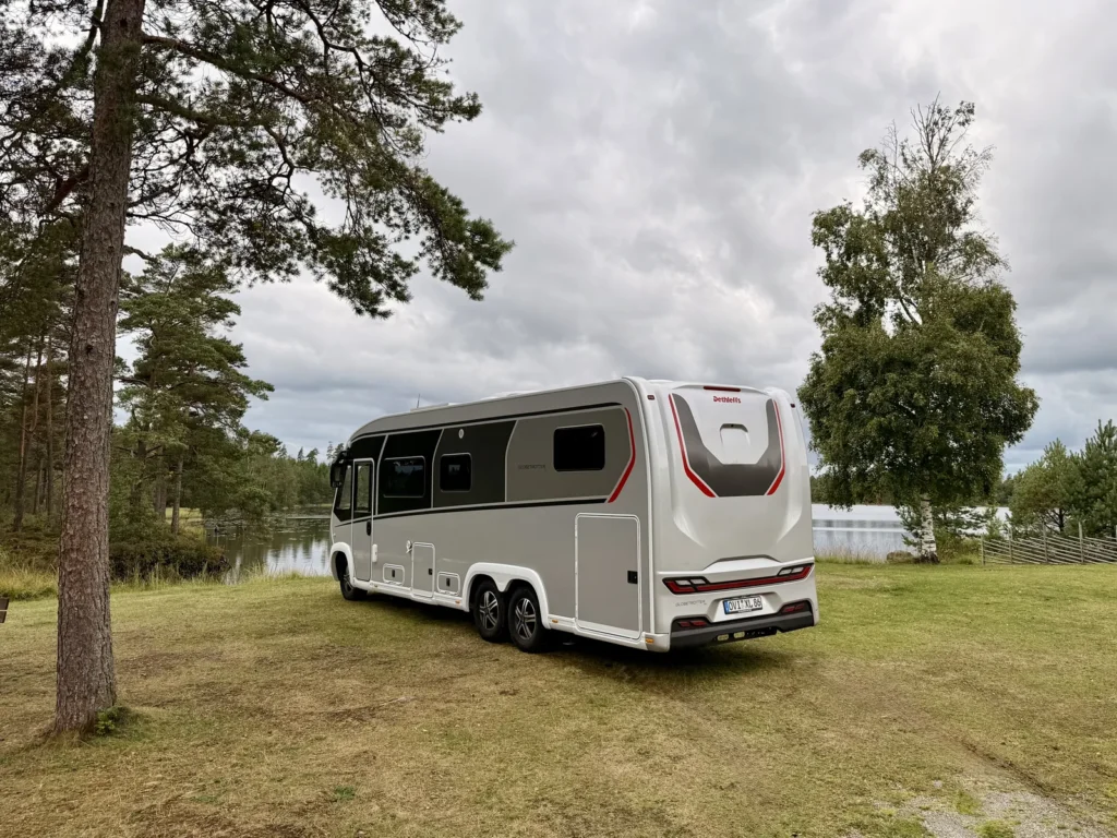 Der Dethleffs Globetrotter XLi steht von hinten fotografiert auf der Wiese am Ufer des Kvarnsjön, links eine Kiefer, im Hintergrund Wald unter Wolkenhimmel.