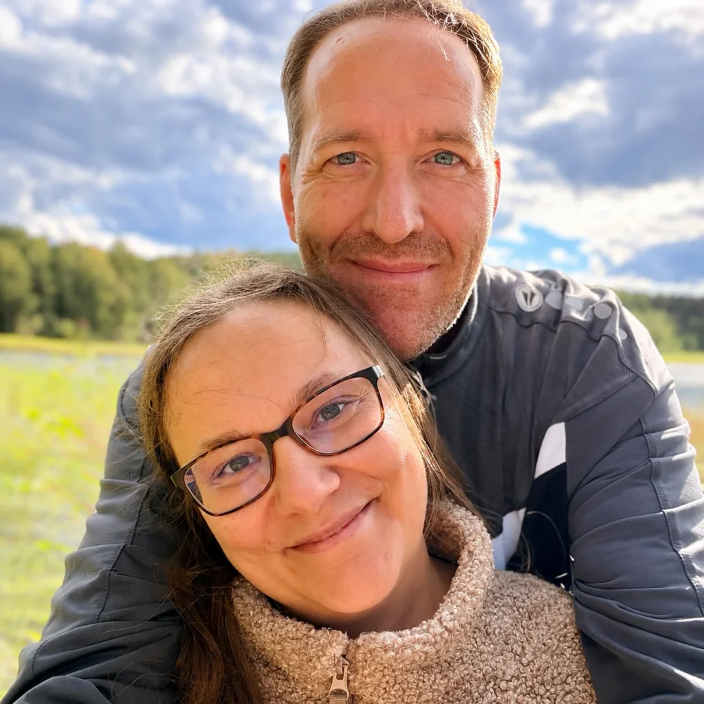 Michael und Maria dicht beieinander im Freien, hinter ihnen eine Wiese, Wald und ein bewölkter Himmel.