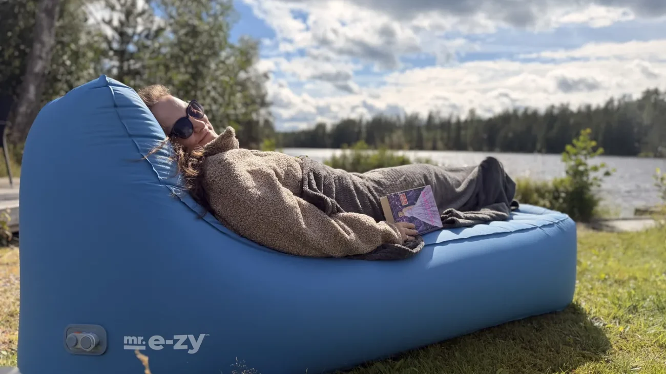 Maria liegt in eine Decke gekuschelt mit einem Buch auf dem petrolblauen mr. e-zy Premium Lounger, im Hintergrund ein See und Wald in Schweden