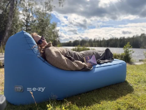 Maria liegt in eine Decke gekuschelt mit einem Buch auf dem petrolblauen mr. e-zy Premium Lounger, im Hintergrund ein See und Wald in Schweden