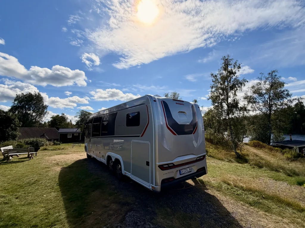 Unser Wohnmobil Luna von schräg hinten auf dem Stellplatz, die hintere Doppelachse ist gut zu sehen, dahinter der See und rote Holzhäuser.