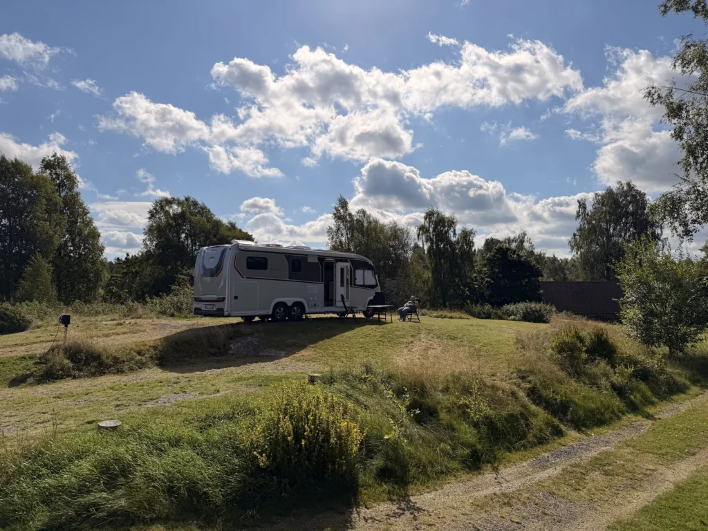Unser Wohnmobil steht auf der erhöhten Wiese am River Lodge Camping, davor Campingtisch und Stühle, ringsum Birken und Wiese.