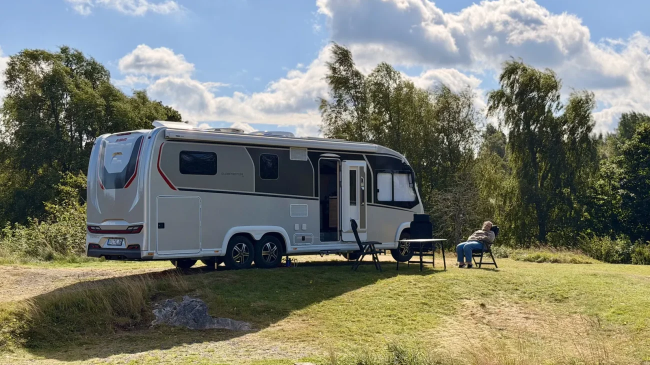 Unser Wohnmobil Luna steht auf der Wiese über dem Wasser am River Lodge Camping in Nyfors, daneben Campingtisch und Stühle, im Hintergrund Birken.