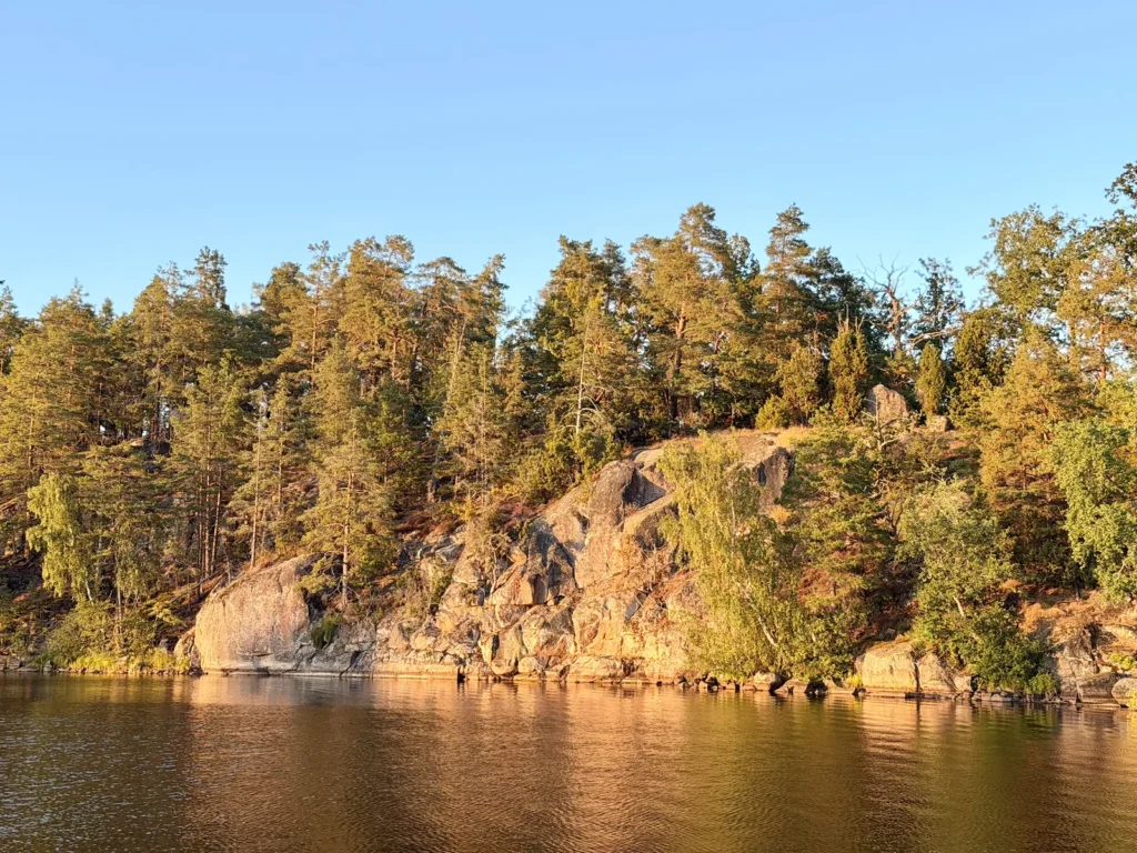 Eine bewaldete Felsklippe am Åsunden leuchtet in der tief stehenden Abendsonne, davor liegt das ruhige Wasser des Sees.
