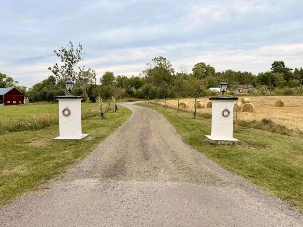Kiesweg zwischen zwei weißen Torpfeilern mit Laternen, links eine rote Scheune, rechts ein abgeerntetes Feld mit Strohballen.