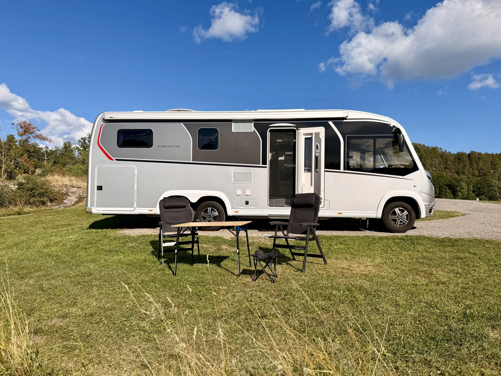 Luna steht seitlich auf dem verdichteten Schotter, davor auf dem Rasen ein Falttisch mit zwei Campingstühlen und einem Hocker.