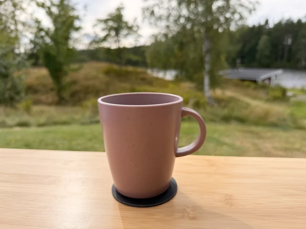 Ein rosafarbener Kaffeebecher steht auf einem schwarzen Magnetpad auf dem Campingtisch, dahinter Wiese, Bäume und ein See.