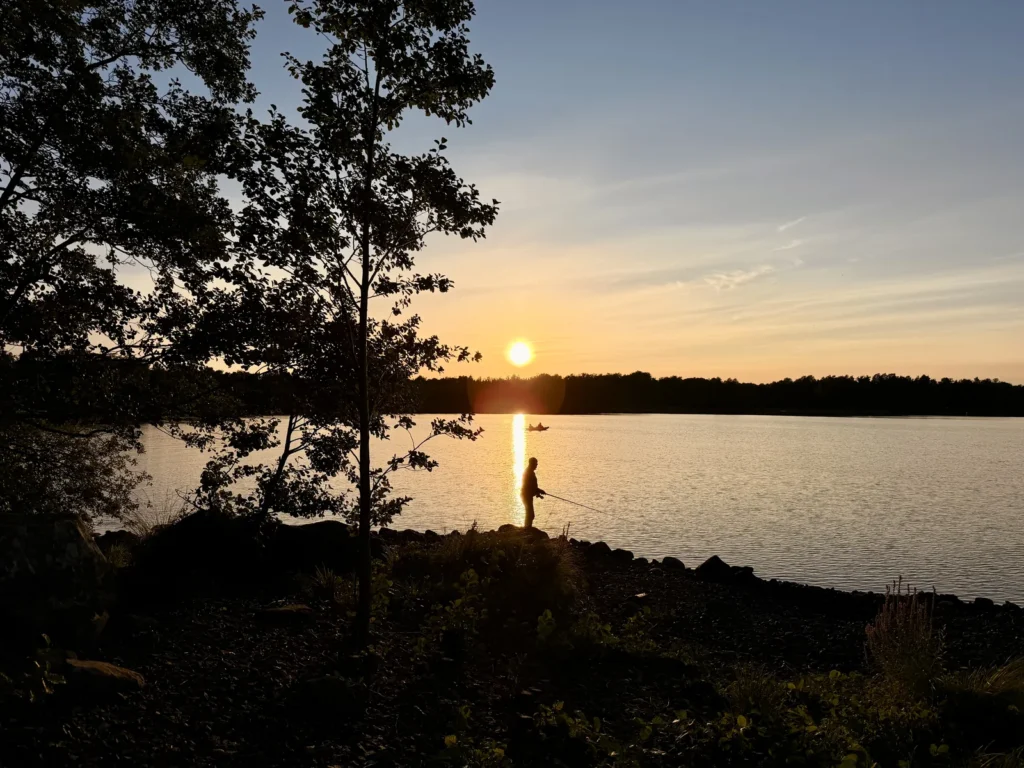 Ein Angler steht als Silhouette am Ufer des Möckeln, die Sonne geht hinter dem Wald unter, im Wasser ein Kajak.