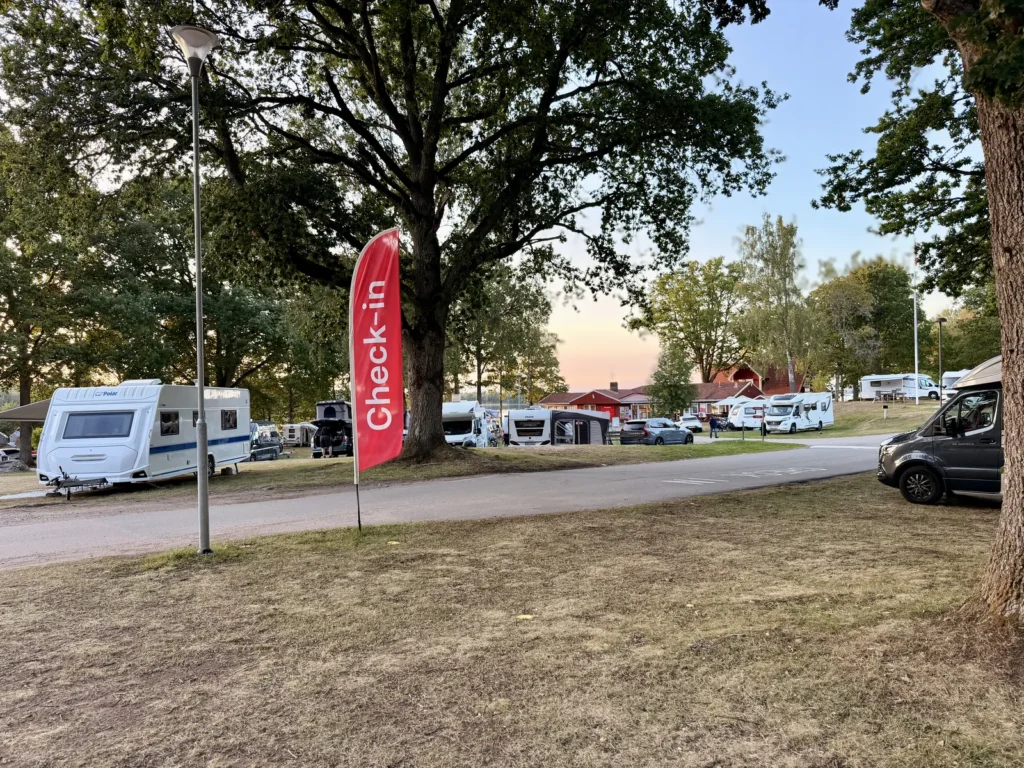 Rote Check-in-Fahne unter einer großen Eiche, dahinter Wohnwagen, Wohnmobile und das rote Rezeptionsgebäude im Abendlicht.