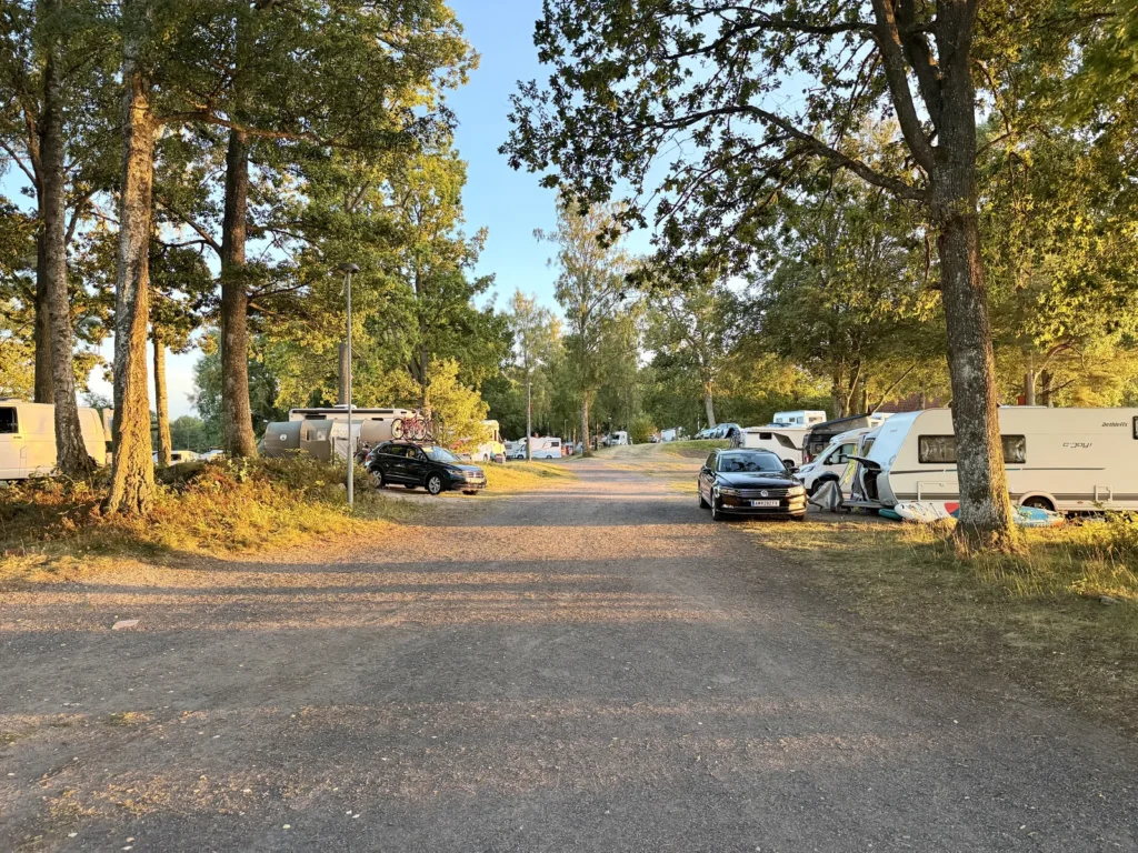 Breiter Schotterweg zwischen den Stellplätzen, links und rechts stehen Wohnwagen und Wohnmobile dicht an dicht unter Eichen.