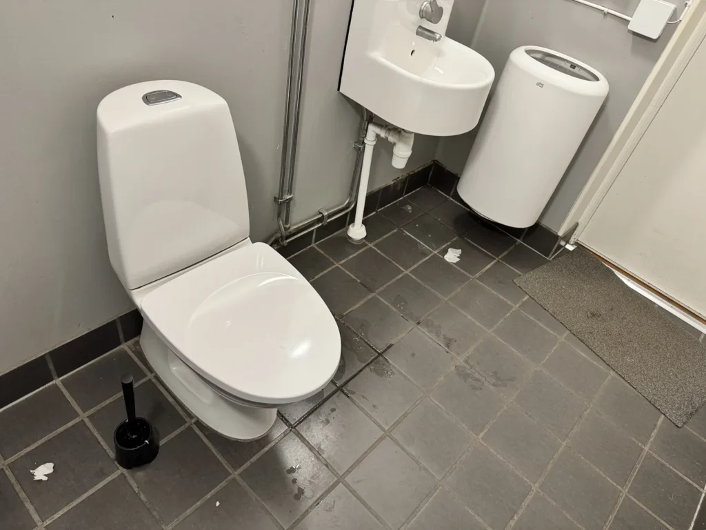 Toilette und Waschbecken in einem Sanitärraum, auf dem gefliesten Boden liegen Papierreste und Pfützen.