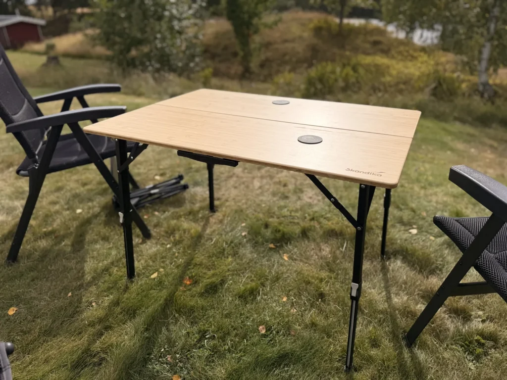 Aufgebauter Skandika Tobro Falttisch mit Bambusplatte auf einer Wiese, daneben zwei Campingstühle, im Hintergrund ein rotes Holzhaus in Schweden