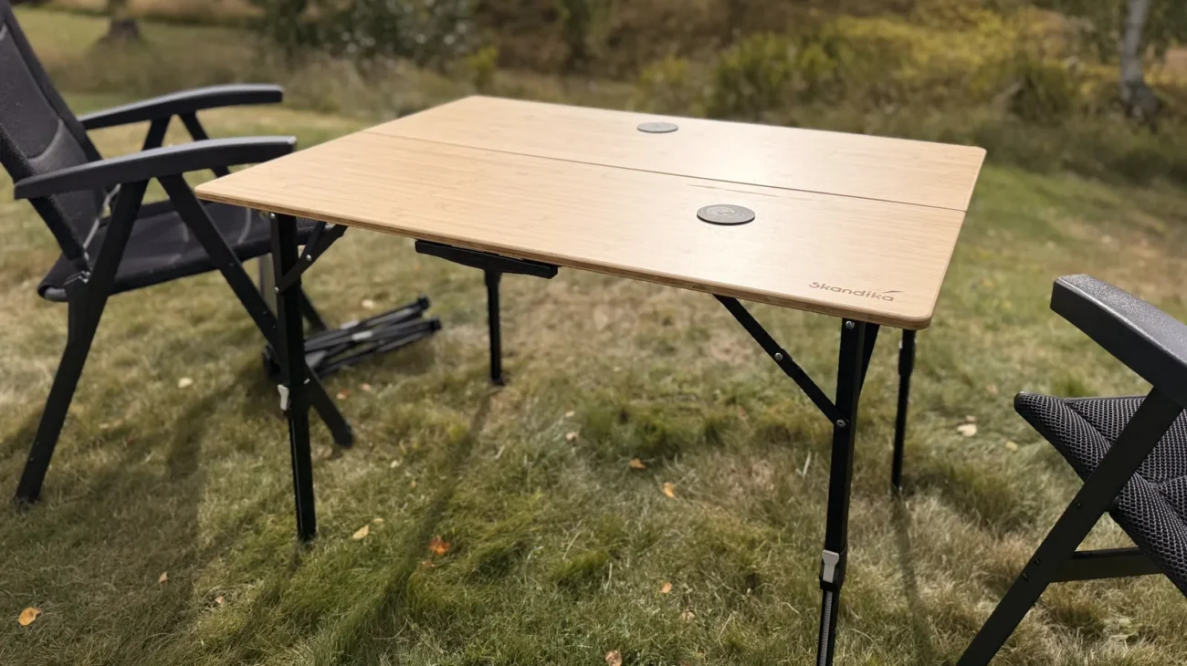 Aufgebauter Skandika Tobro Falttisch mit Bambusplatte auf einer Wiese, daneben zwei Campingstühle, im Hintergrund ein rotes Holzhaus in Schweden