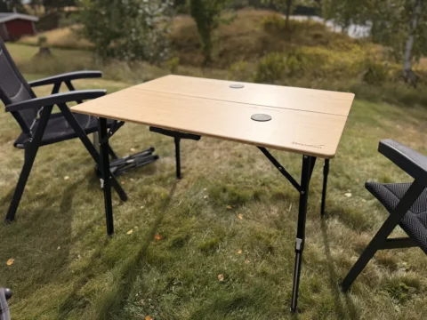 Aufgebauter Skandika Tobro Falttisch mit Bambusplatte auf einer Wiese, daneben zwei Campingstühle, im Hintergrund ein rotes Holzhaus in Schweden