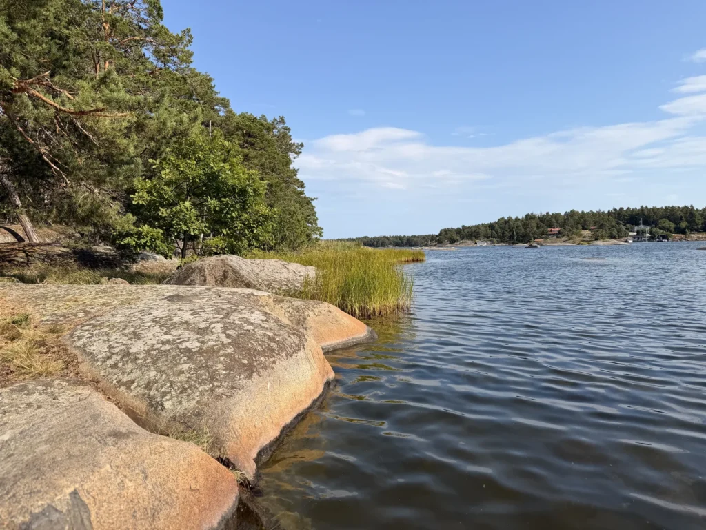Rundgeschliffene Felsen am Ufer, davor Schilf im flachen Wasser, am gegenüberliegenden Ufer rote Holzhäuser.
