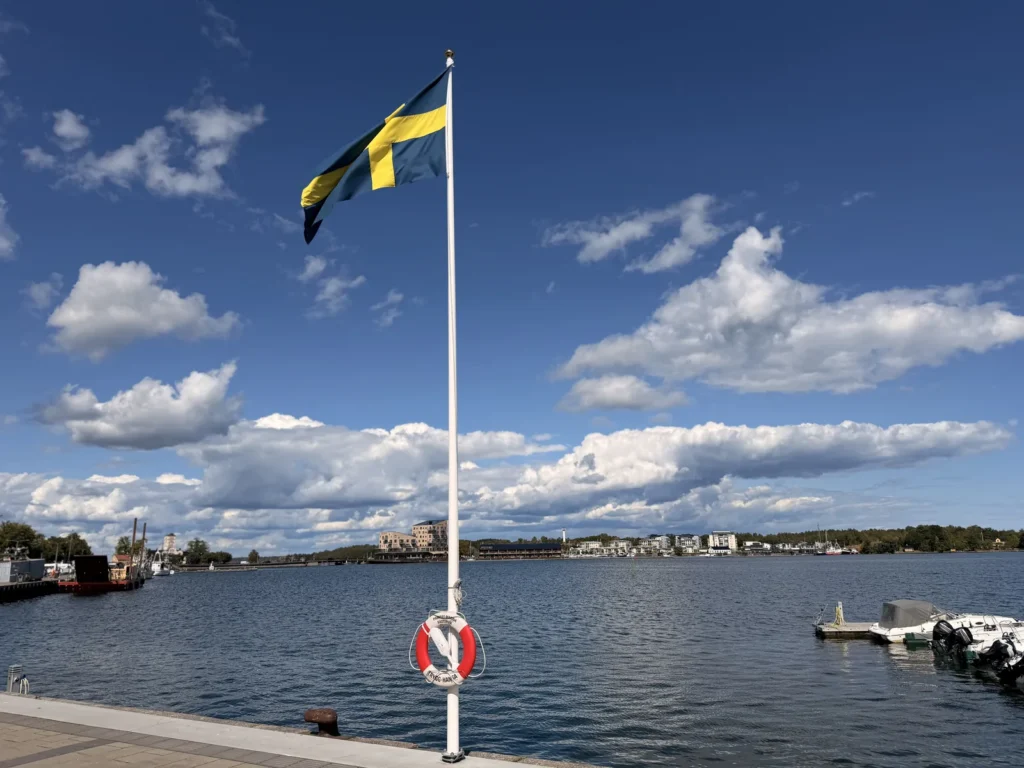 Eine schwedische Flagge weht an einem weißen Mast am Kai, dahinter das Wasser und die Häuser von Västervik unter einem Himmel mit Quellwolken.