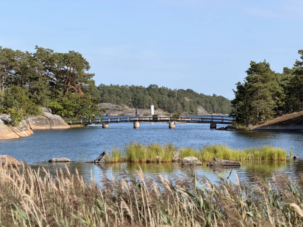 Eine niedrige Holzbrücke führt über einen schmalen Meeresarm, im Vordergrund Schilf und Felsen.