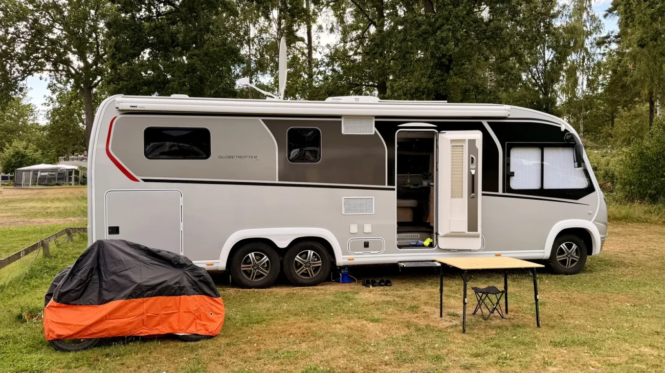 Der weiße Dethleffs Globetrotter XLi steht mit offener Aufbautür auf einer Wiese unter hohen Laubbäumen, davor ein Campingtisch und ein abgedecktes Fahrrad.