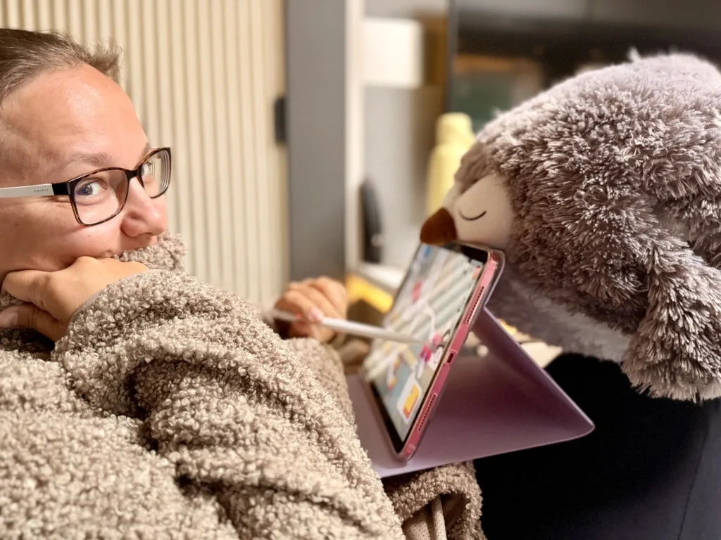 Maria liegt im Wohnmobil eingemummelt in einem Teddyfell-Pullover, daneben ein Tablet mit einer Zeichen-App und ein Plüschtier.