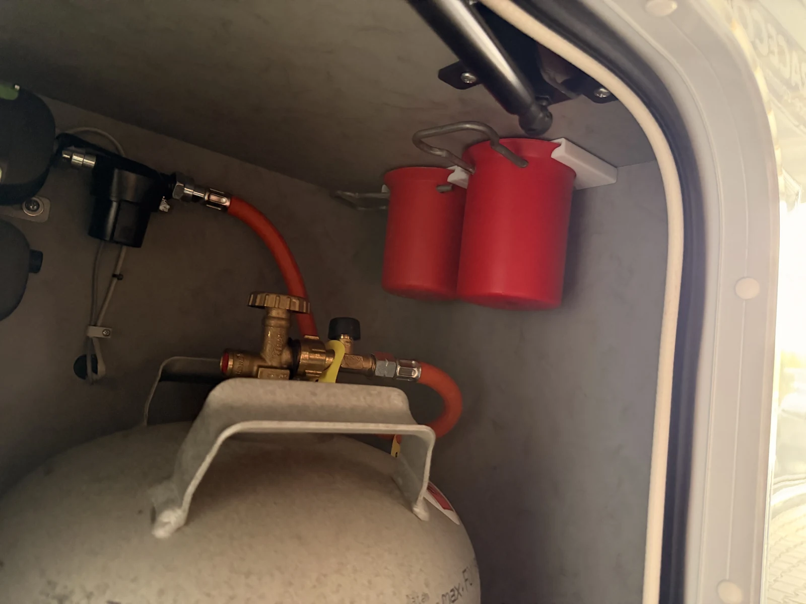 Zwei rote Gasflaschendeckel stecken in einem weißen 3D-gedruckten Halter, der oben im Gaskasten klebt