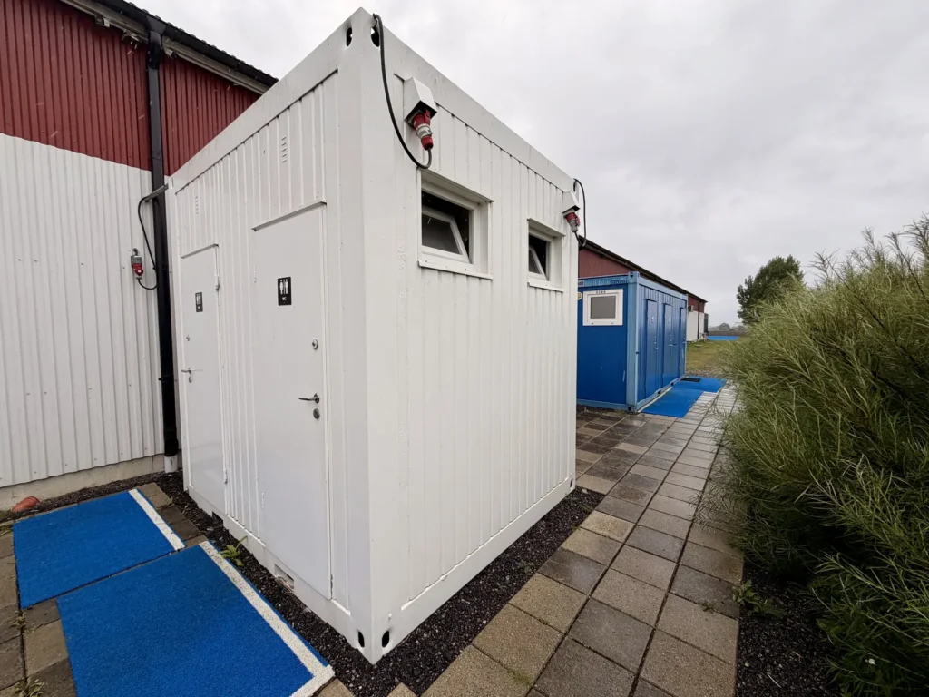 Weiße und blaue Sanitärcontainer mit WC-Schildern stehen an einer roten Halle, davor blaue Kunstrasenmatten und ein gepflasterter Weg.