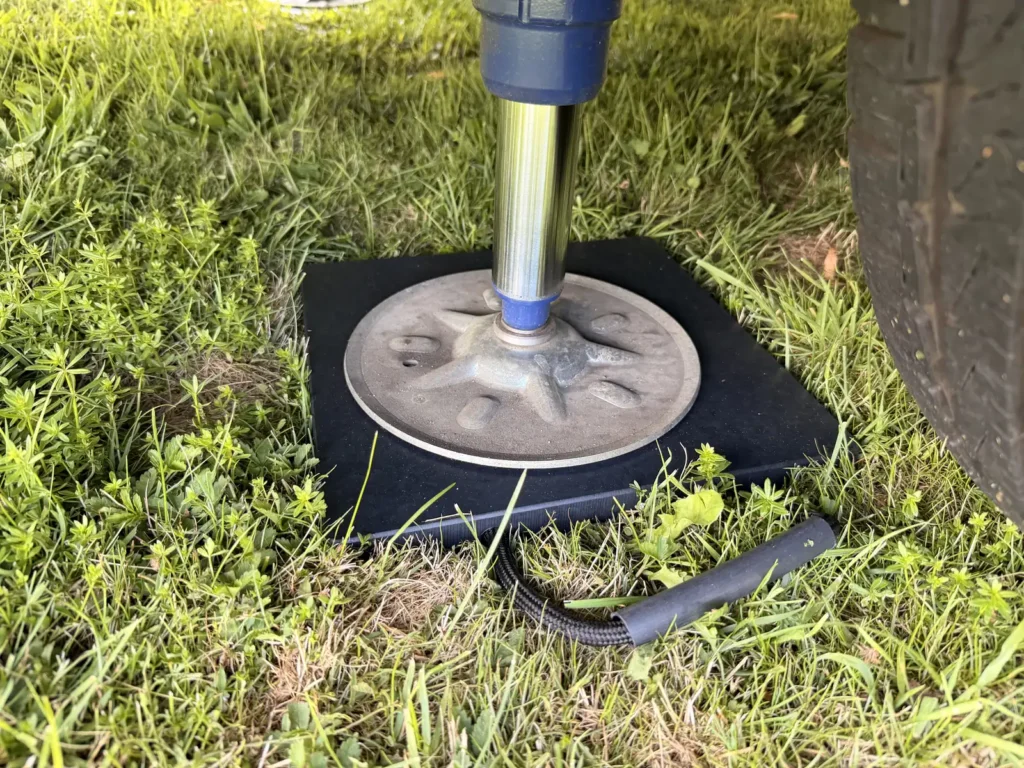 Ausgefahrene Hubstütze des Wohnmobils steht auf einer schwarzen Stützplatte im Gras, daneben der Reifen
