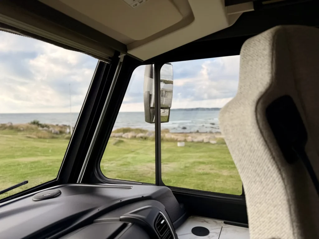 Blick vom Beifahrersitz durch die Frontscheibe über die Wiese auf das Steinufer und das Meer, im Außenspiegel spiegelt sich der Nachbarplatz.