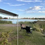 Maria sitzt unter der ausgefahrenen Markise von Luna im Campingstuhl am Tisch, dahinter Schilf und der Binnensee bei Bergkvara