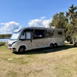 Der Dethleffs Globetrotter XLi steht auf der Wiese von Stellplatz 199, links das Wasser mit Schilfgürtel, rechts hohe Wacholderbäume.