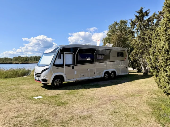 Der Dethleffs Globetrotter XLi steht auf der Wiese von Stellplatz 199, links das Wasser mit Schilfgürtel, rechts hohe Wacholderbäume.