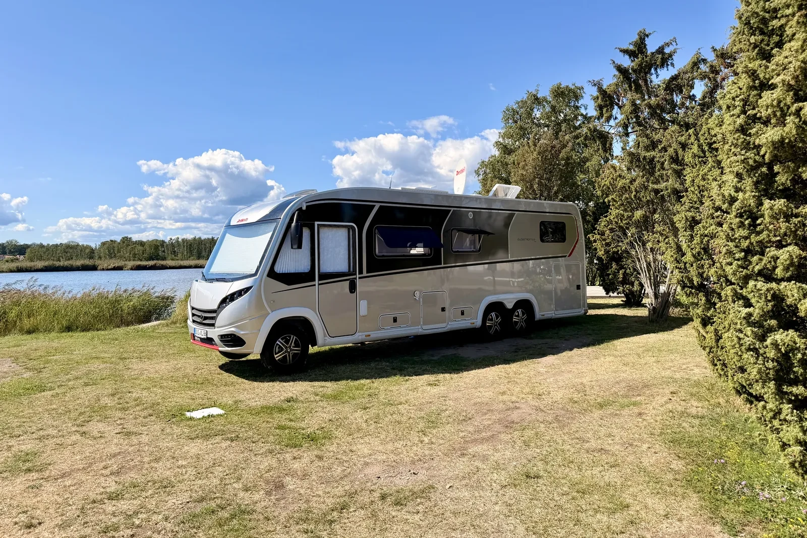 Der Dethleffs Globetrotter XLi steht auf der Wiese von Stellplatz 199, links das Wasser mit Schilfgürtel, rechts hohe Wacholderbäume.