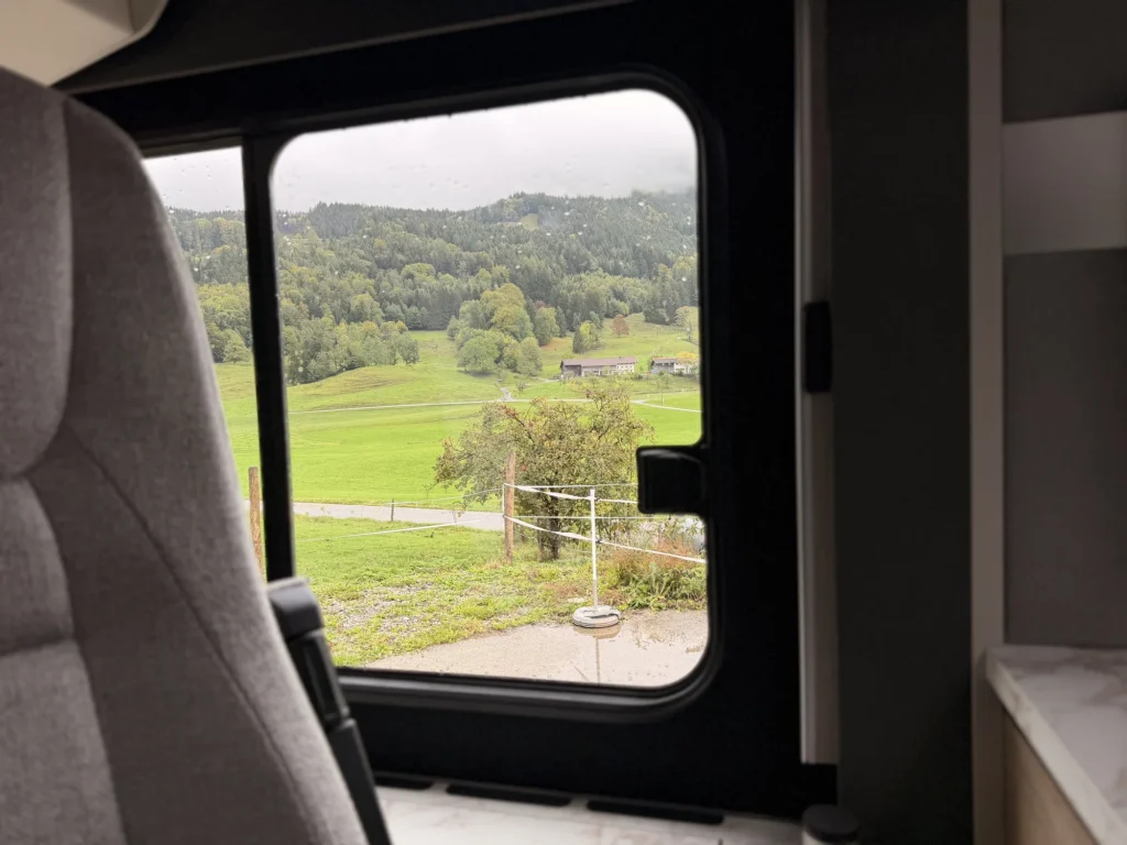 Blick aus dem Seitenfenster des Wohnmobils auf nasse Wiesen, Bäume und bewaldete Hänge unter tiefen Wolken