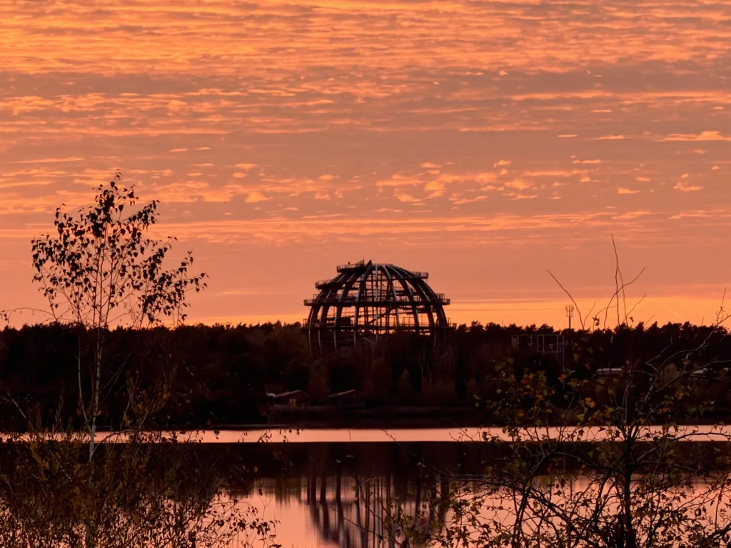 Die große Holzkugel am Steinberger See als Silhouette vor orangem Abendhimmel, im Vordergrund das Wasser und Büsche