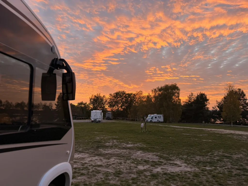 Orange leuchtende Wolken beim Sonnenuntergang über dem Stellplatz am Steinberger See, links Lunas Außenspiegel, hinten drei Wohnmobile vor Bäumen