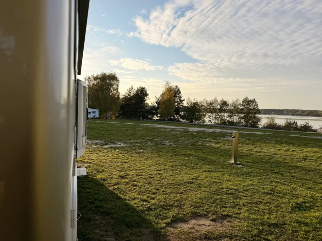 Blick an Lunas Seitenwand vorbei über die Wiese des Stellplatzes mit einer Stromsäule, dahinter Birken und das Wasser des Steinberger Sees